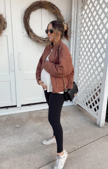 A pregnant woman wearing a quilted brown jacket over a white top, paired with black leggings and sneakers, perfect for casual outings.