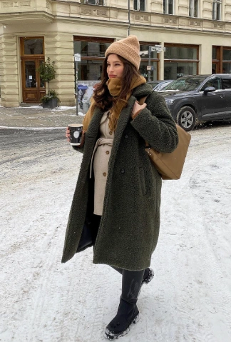 Long olive green maternity coat, layered over a beige top, paired with black pants and a knit beanie, perfect for casual winter outings.
