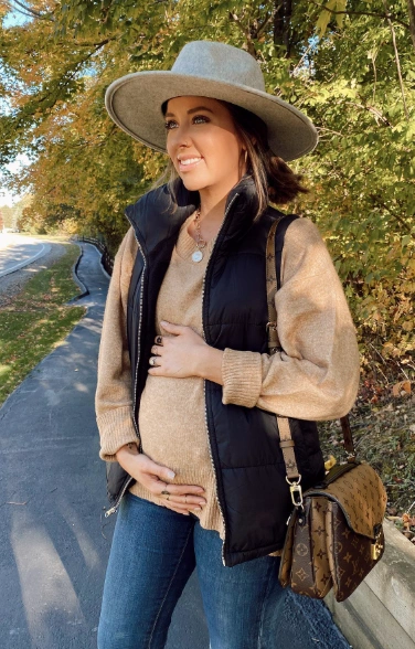 Maternity outfit featuring a beige knit sweater, black puffer vest, and blue jeans. Ideal for casual autumn outings.