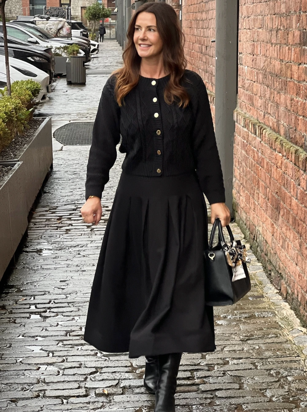 Black ribbed cardigan paired with a flowing midi skirt, sleek ankle boots, and a structured handbag on a wet cobblestone street.