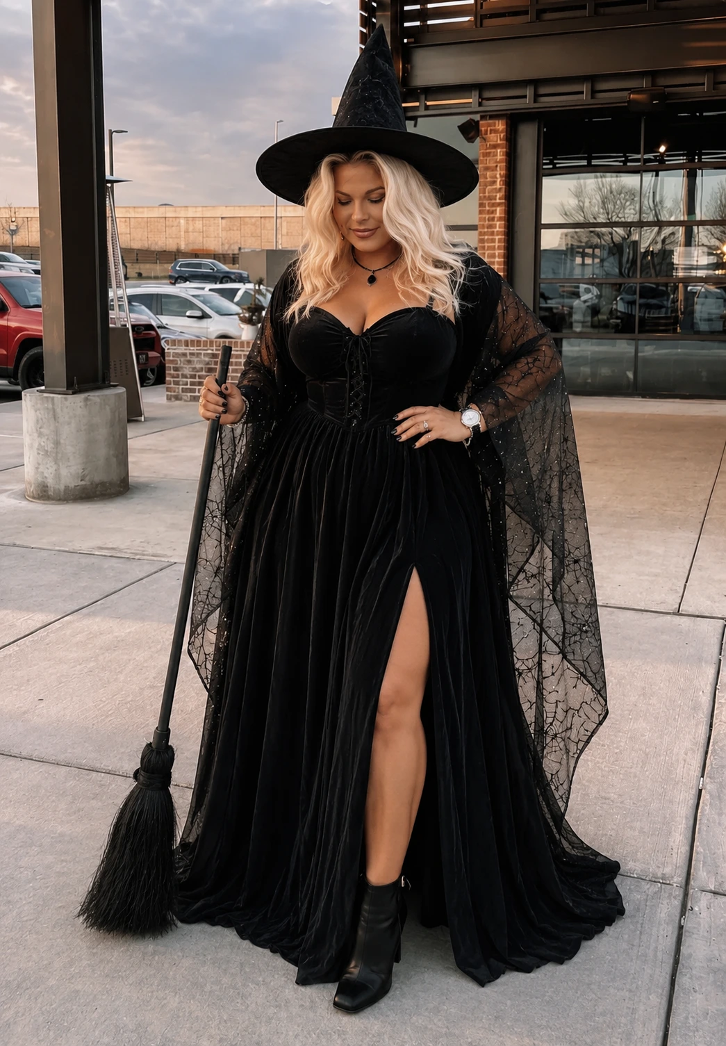 Blonde woman in a black velvet witch costume, pointed hat, sheer cape sleeves, holding a broom outdoors.