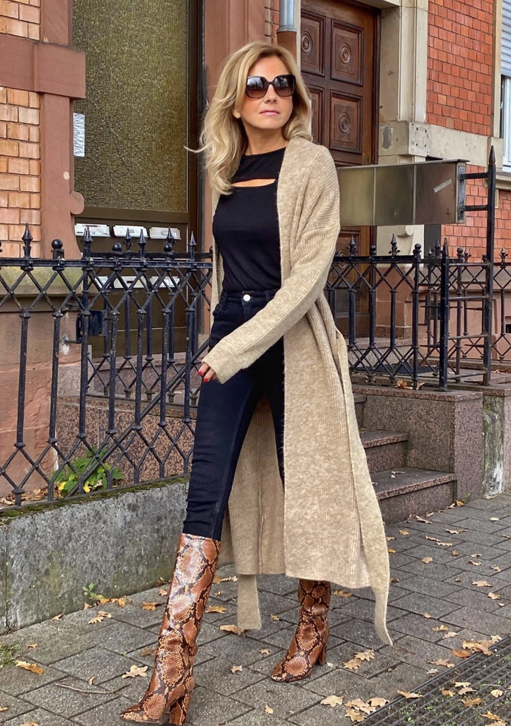 Long beige cardigan layered over a black cutout top, dark skinny jeans, and pointed snakeskin boots on a city sidewalk.