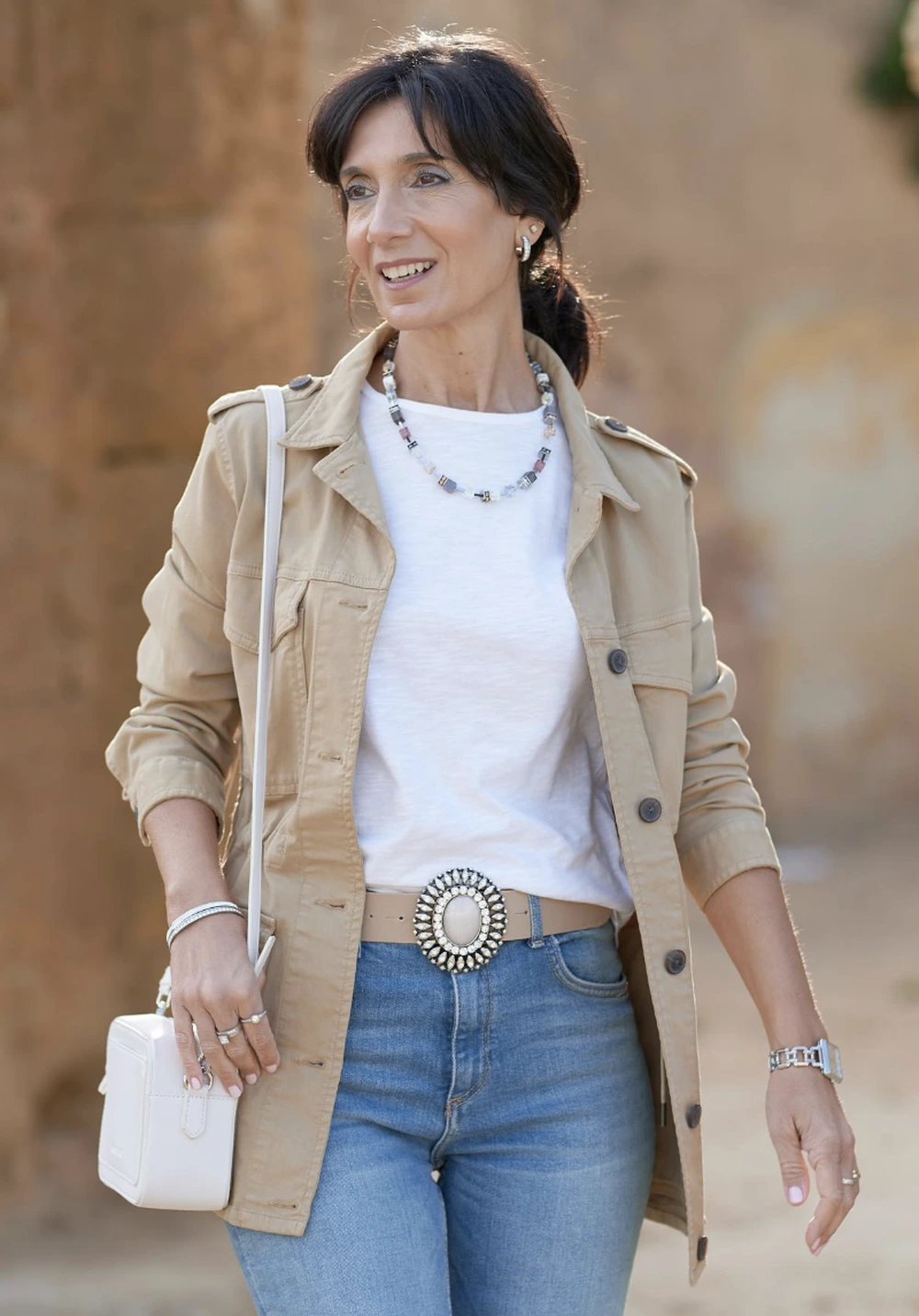 Beige utility jacket over white tee, straight blue jeans, statement oval belt, white crossbody bag, and silver jewelry.