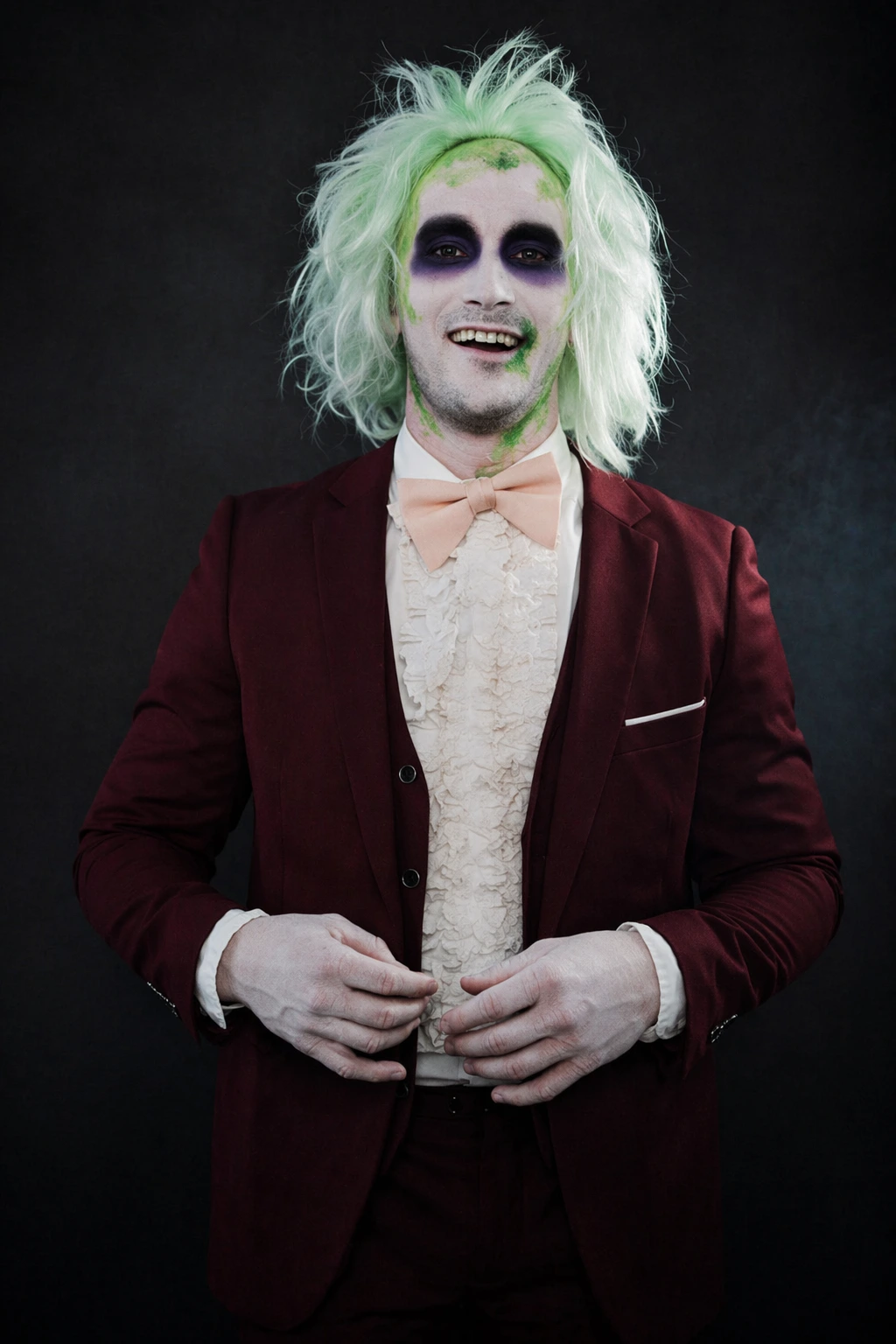 Pale-faced man with messy mint hair, dark eye makeup, burgundy blazer, peach bow tie, and ruffled shirt.