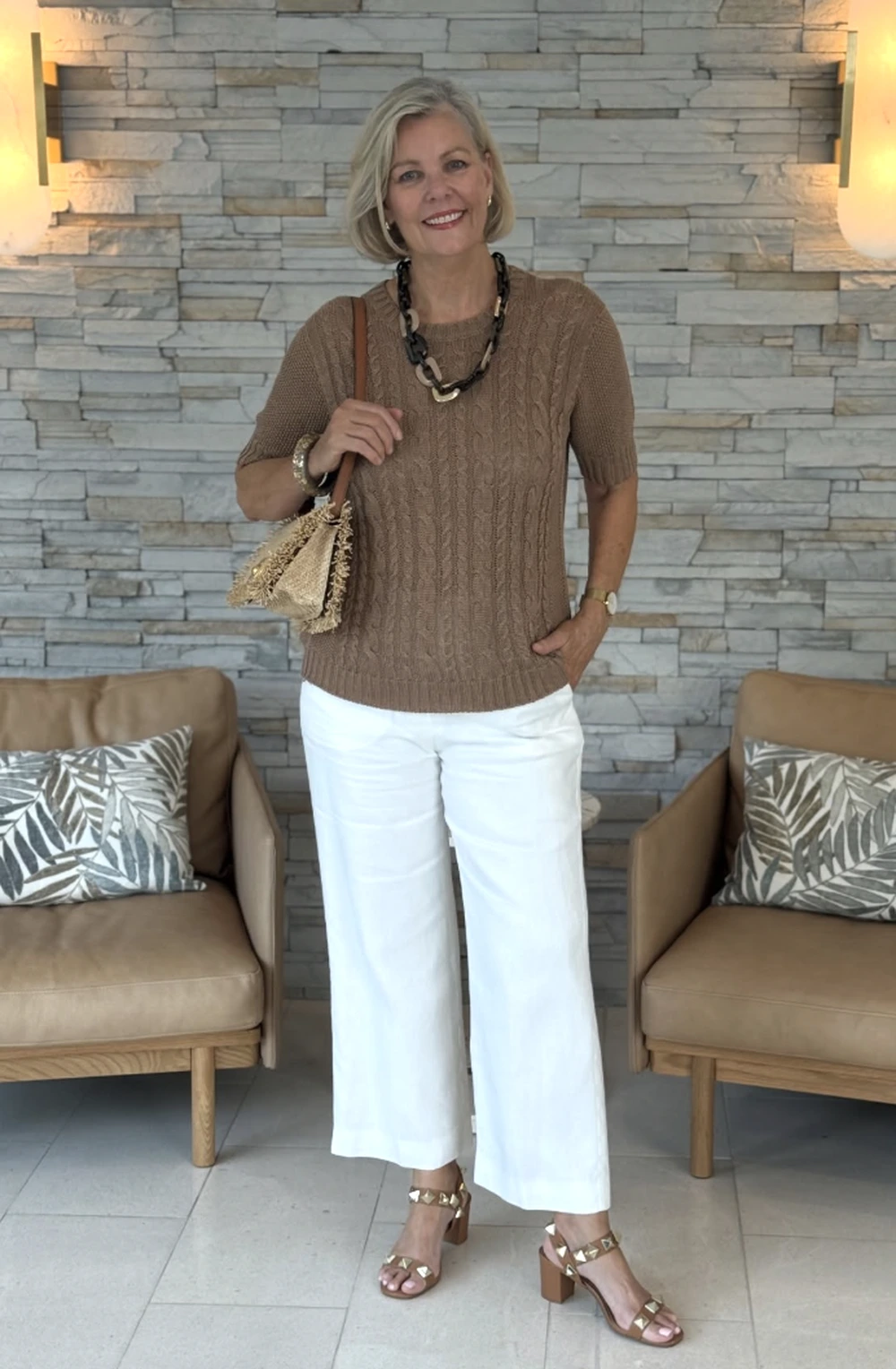 Woman in brown cable-knit sweater, white wide-leg trousers, statement necklace, and strappy heels in a living room.