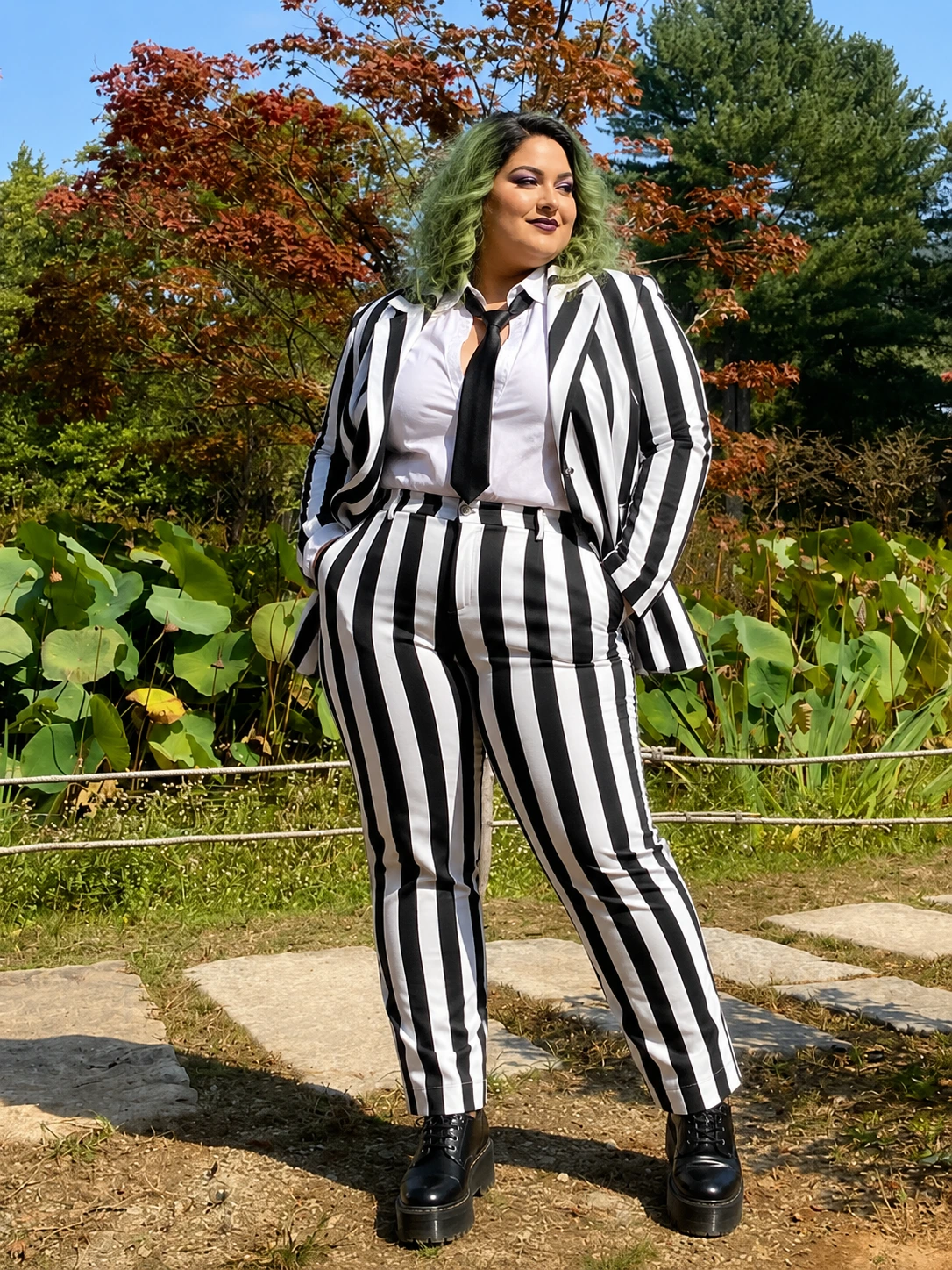 Person in black-and-white striped suit with tie, green hair, and platform shoes standing in a garden.