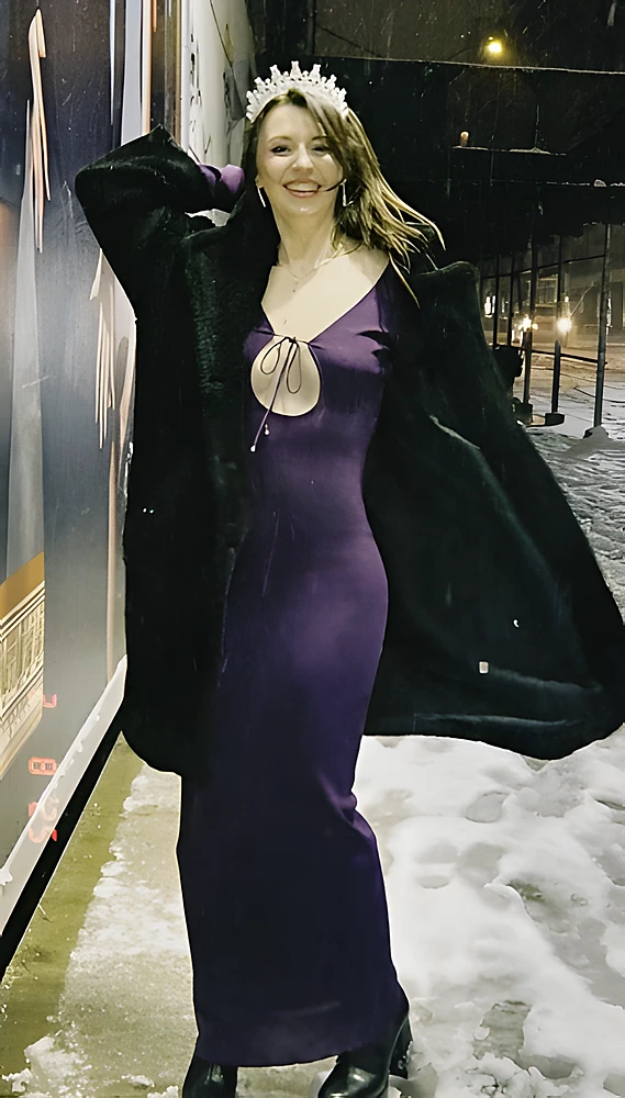 A woman wears a long purple dress with a front cutout, a black faux fur coat, and a tiara.
