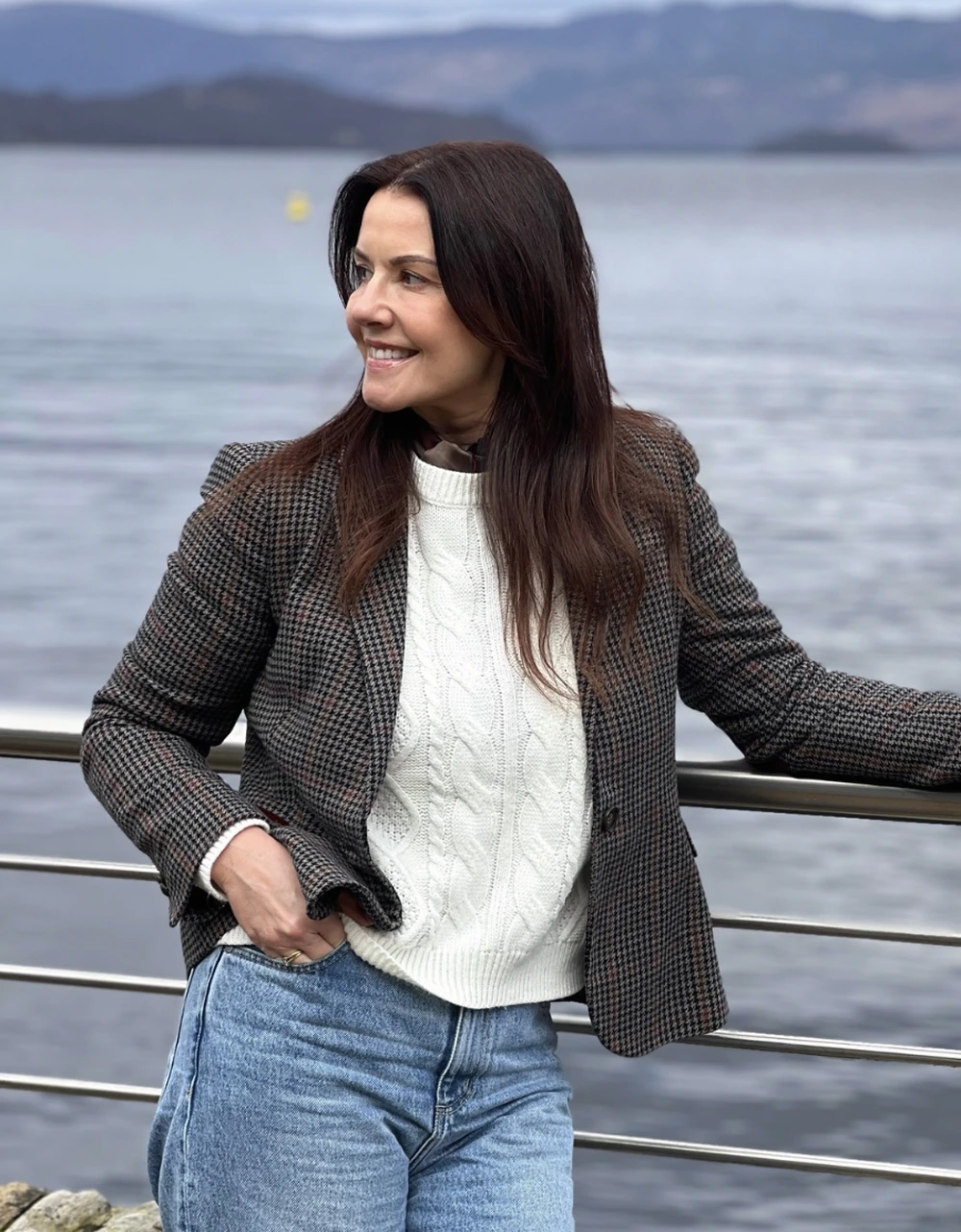 Houndstooth blazer with cream cable-knit sweater, relaxed blue jeans, standing by railing near calm water.