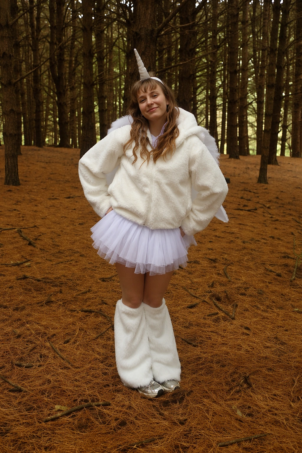 Woman stands in pine forest wearing white fuzzy hoodie, tulle skirt, leg warmers, and unicorn horn.