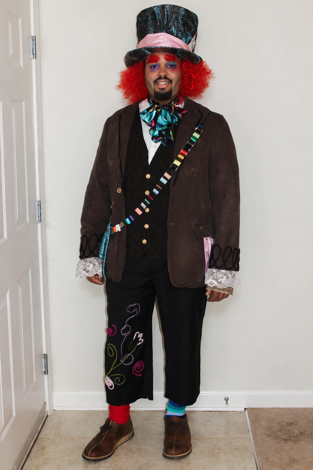 Man with bright red wig, glittery top hat, layered jacket, patterned pants, and playful mismatched accessories.