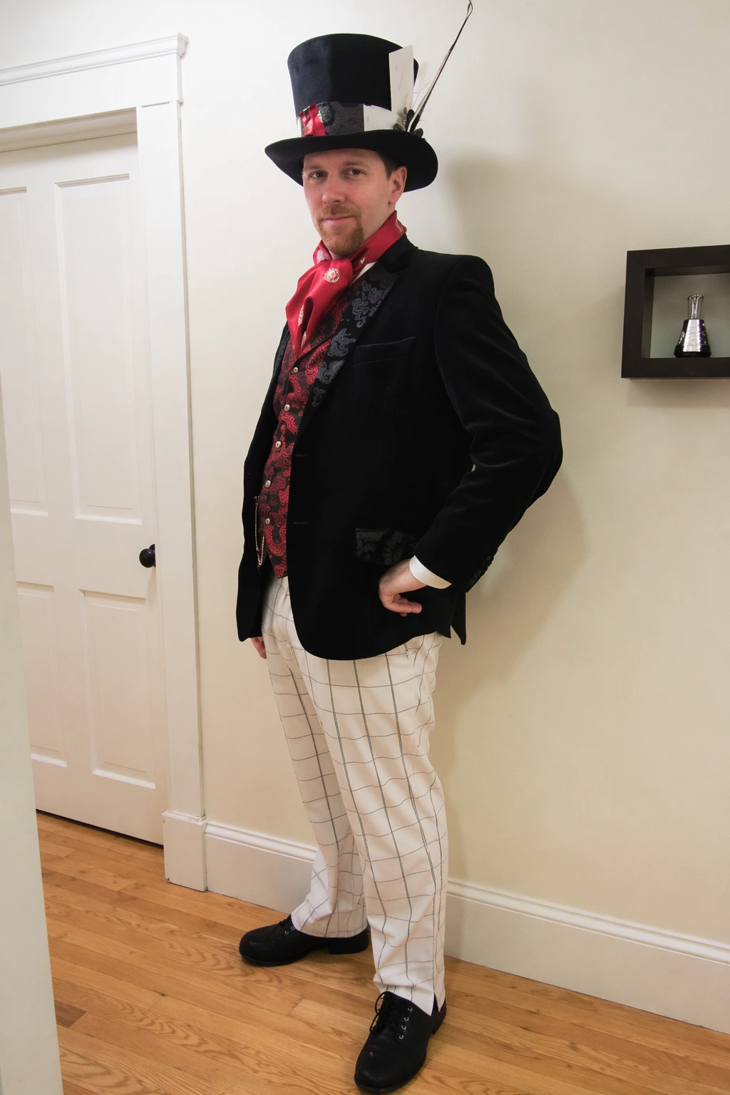 Man posing indoors in a black velvet coat, white check trousers, red scarf, and oversized top hat.