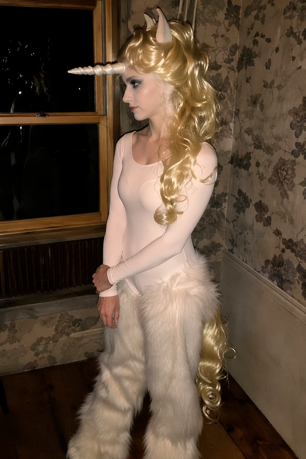 Side-profile of woman in cream unicorn bodysuit, fluffy white fur pants, blonde wig, and horn headpiece.