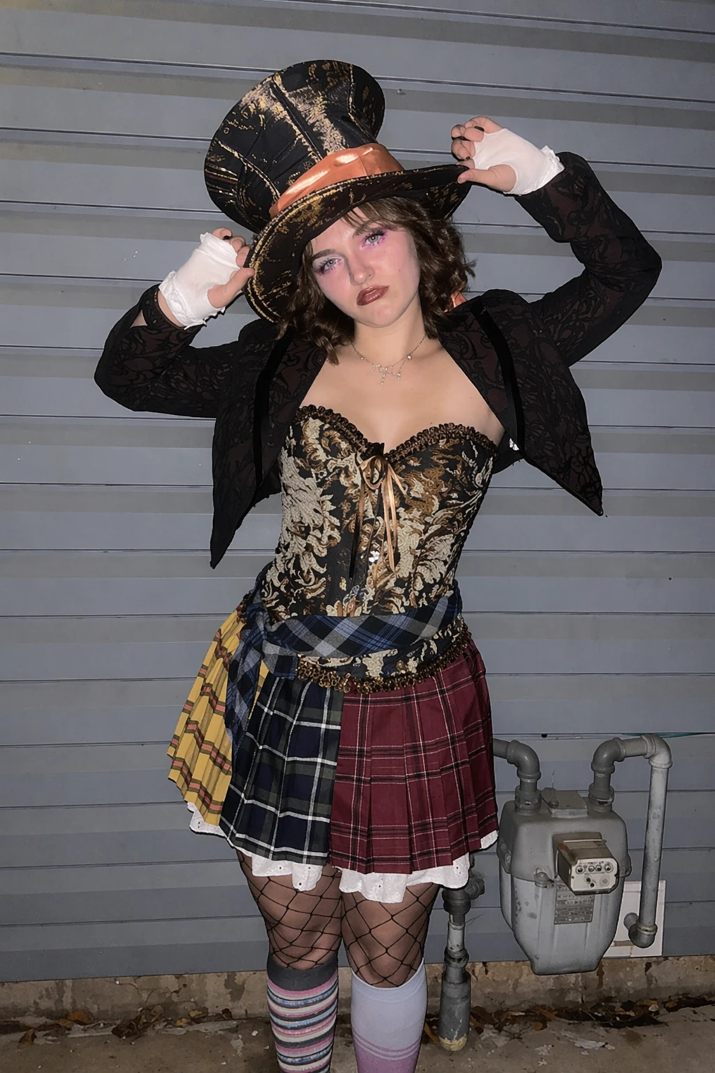 Curly brown hair, black-and-gold top hat, floral corset, cropped lace jacket, tartan skirt, and fishnet tights.