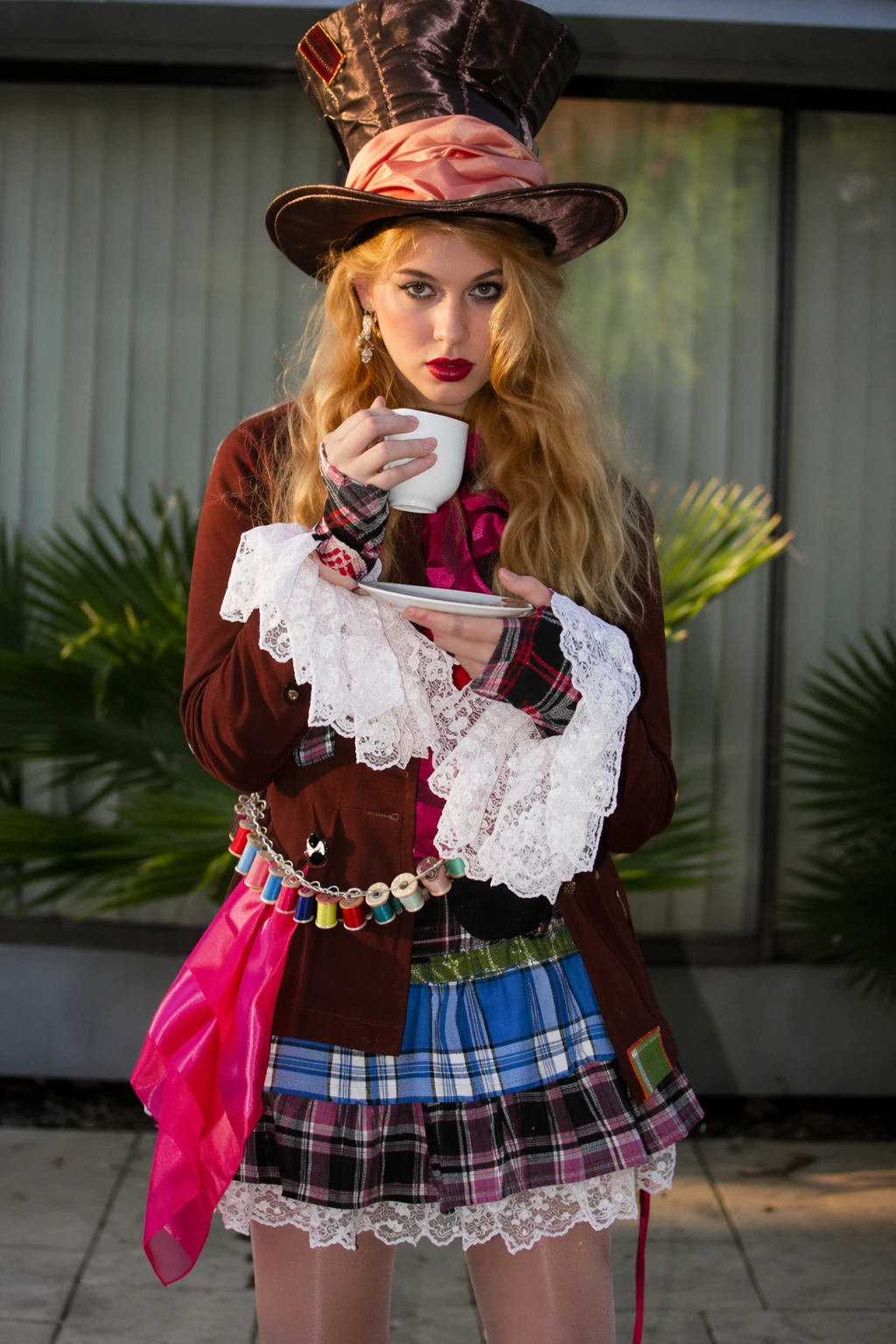 Blonde woman wearing a tall satin hat, lace cuffs, plaid skirt, and colorful sash, sipping from a teacup.