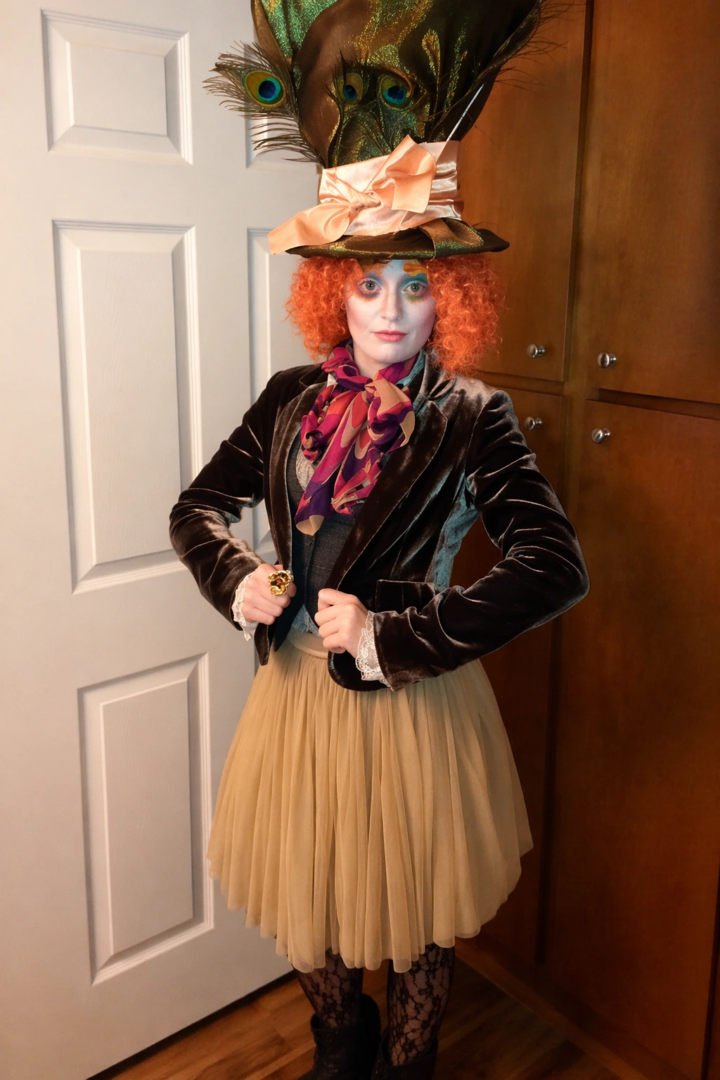 Person with orange curls wearing a towering feathered hat, velvet jacket, pleated skirt, and patterned scarf indoors.