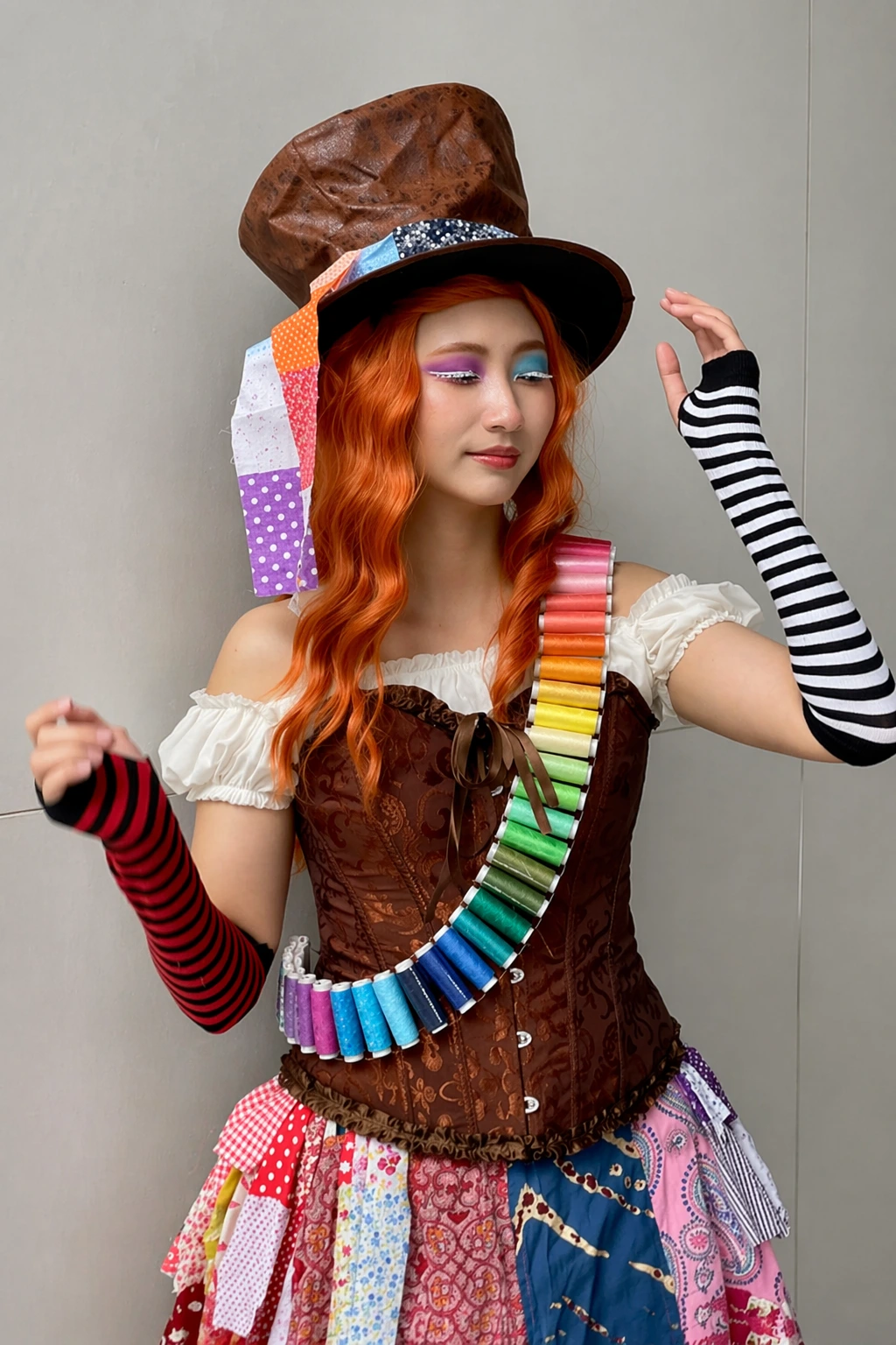 Orange curls, oversized brown top hat, off-shoulder white top, rainbow spool necklace, striped gloves, and patchwork skirt.