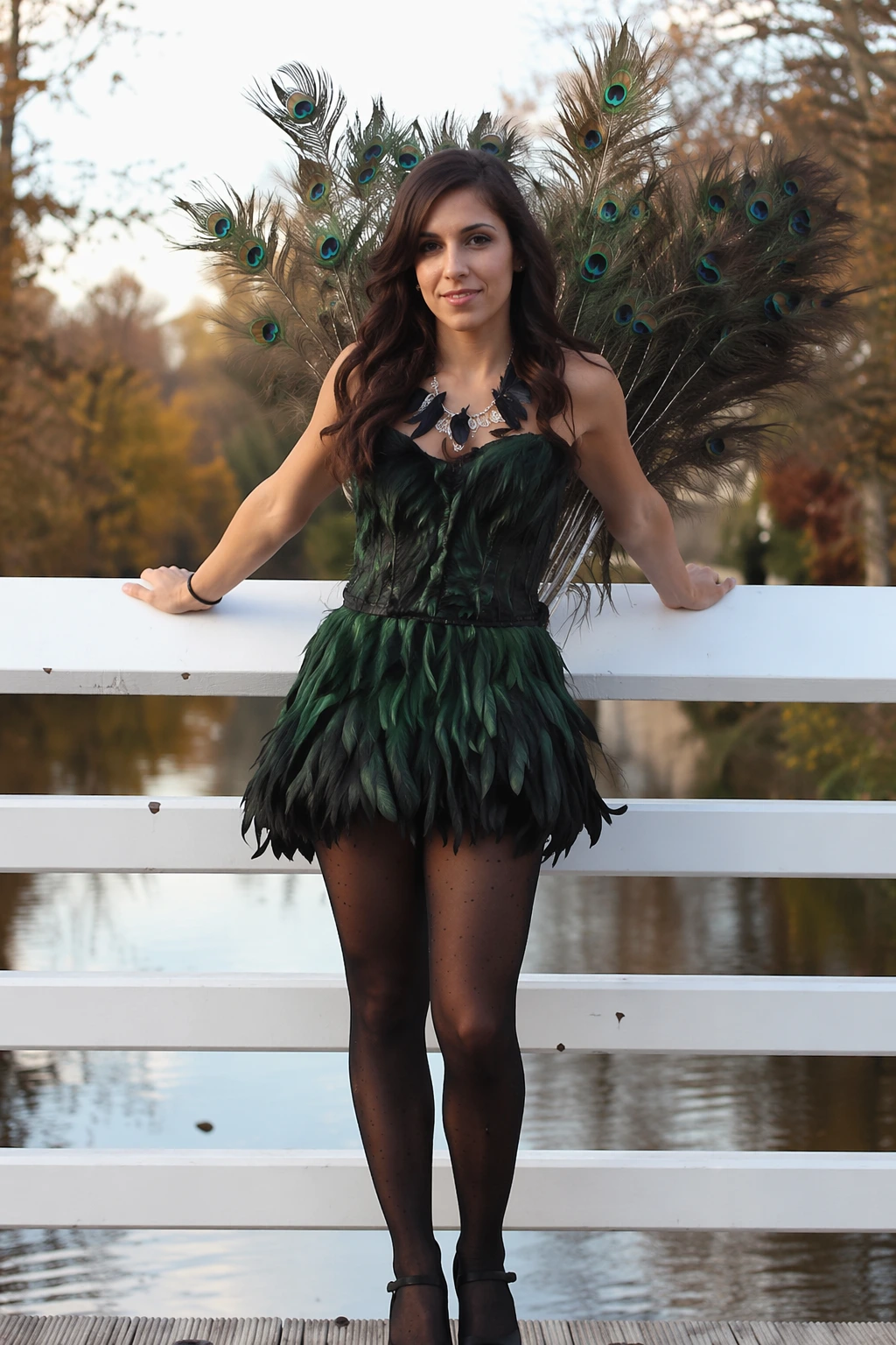 Woman wearing green feather mini dress with peacock tail wings, standing on a white bridge outdoors.