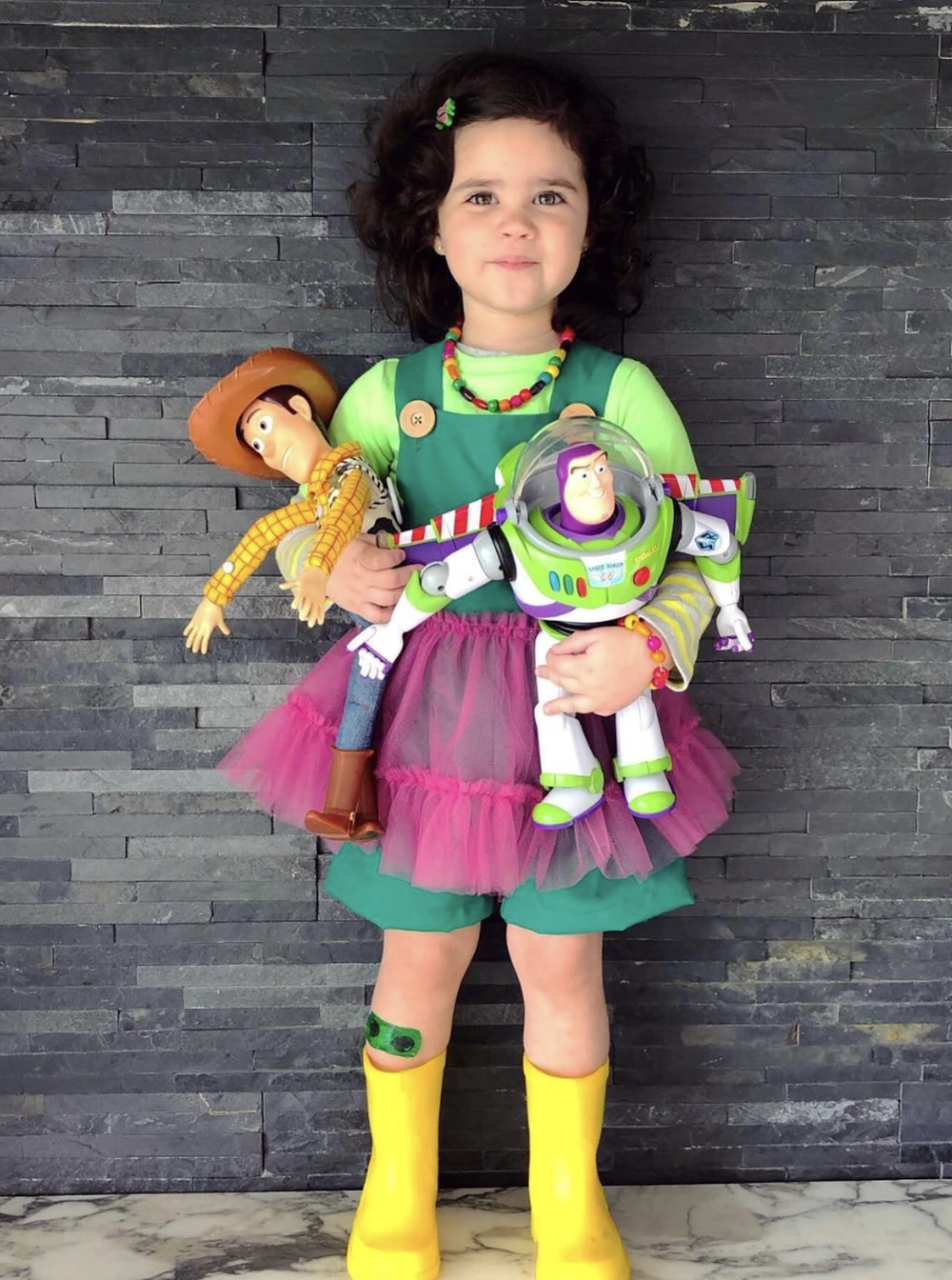 Little girl in a green top, pink tutu, and yellow boots holding Woody and Buzz Lightyear toys.