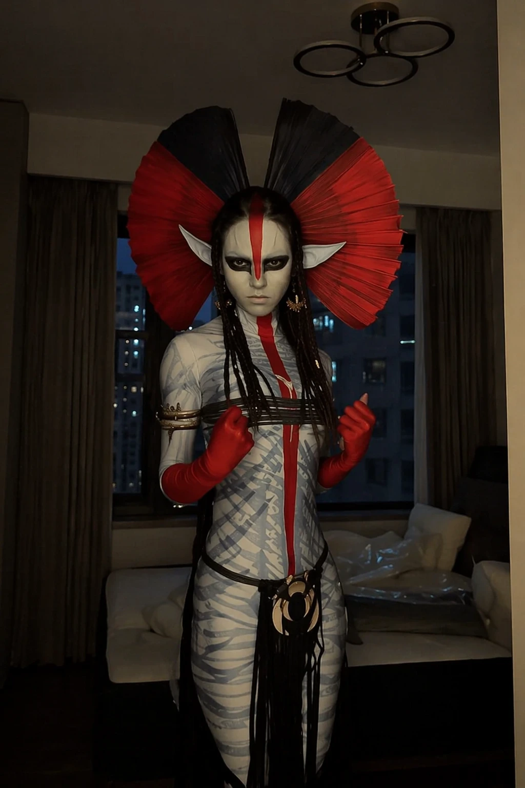 Pale-faced avatar costume with red-black fan headdress, braided hair, red gloves, and patterned body paint.