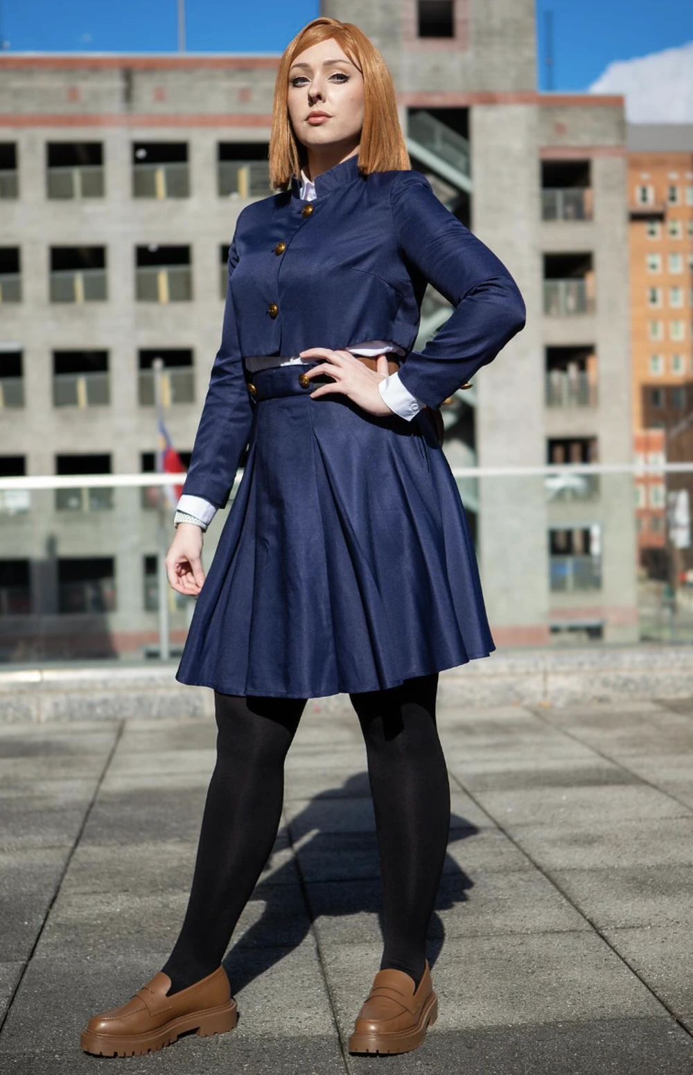 Red-haired cosplayer in navy buttoned jacket and pleated skirt, black tights, and brown loafers on rooftop.