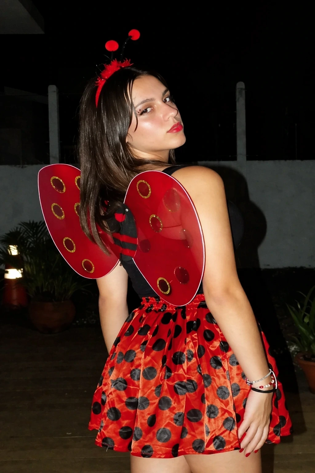 Young woman in a black top and red polka-dot skirt, wearing translucent red wings and antenna headband outdoors.