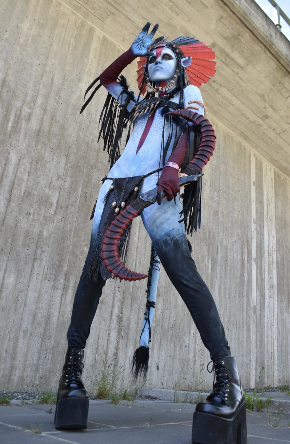 Tall blue avatar in dramatic asymmetrical costume, platform boots, red headdress, long fringe, and curved sculptural accessories.