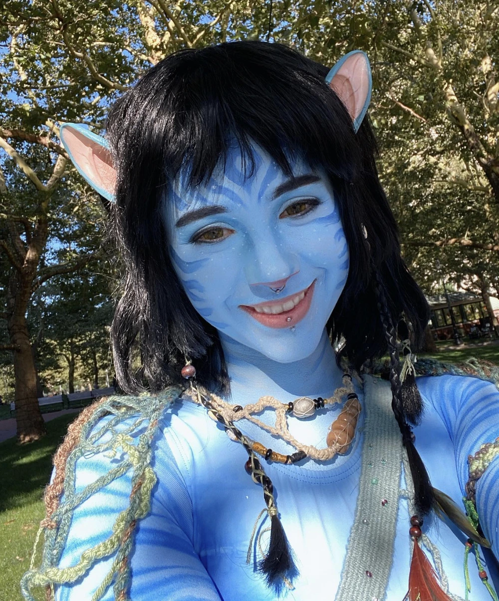 Blue-painted avatar face with black wig, pointed ears, layered bead necklaces, and braided shoulder details outdoors.