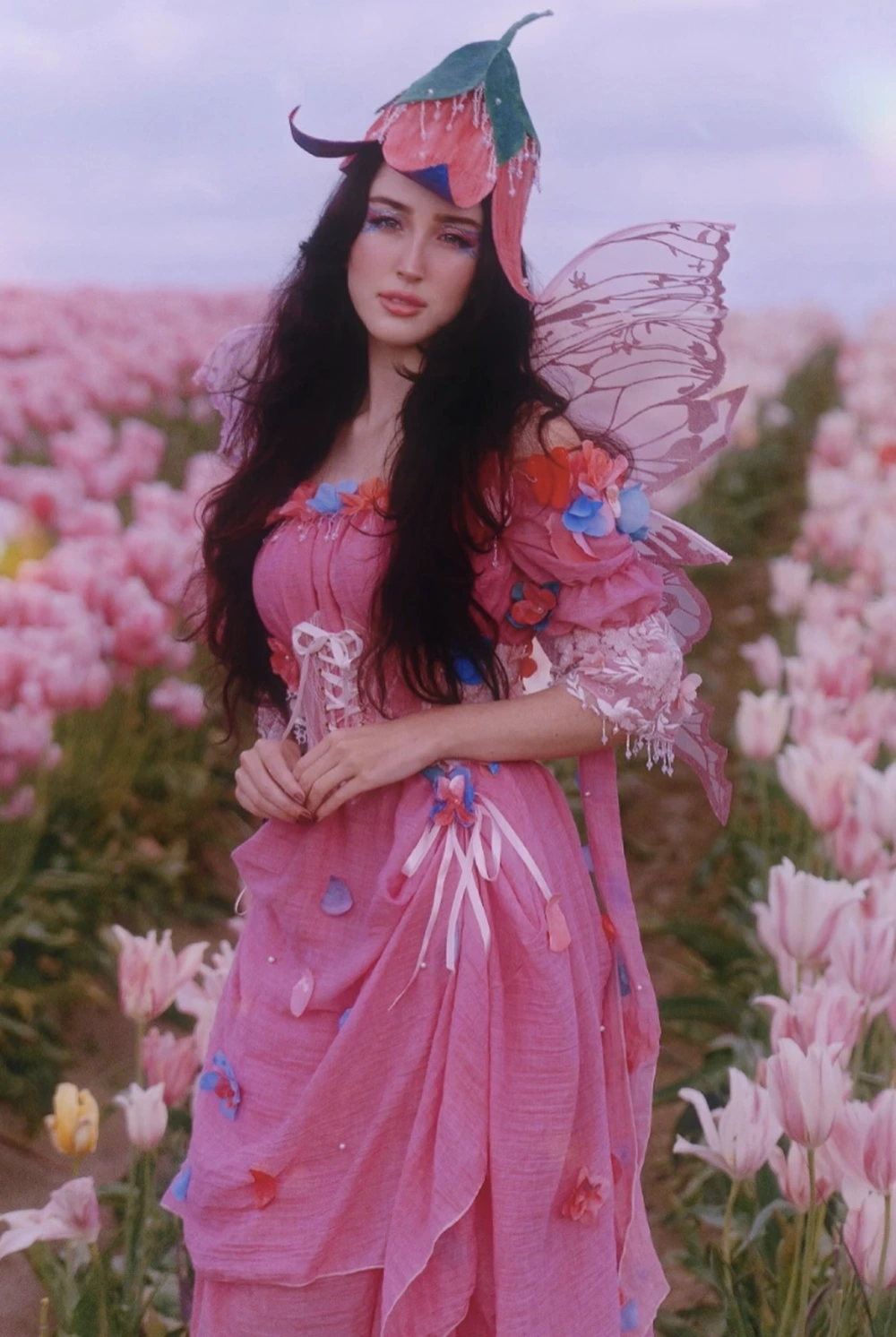 Woman wearing a pink flower fairy dress, petal hat, translucent wings, and standing among tall pink blossoms.