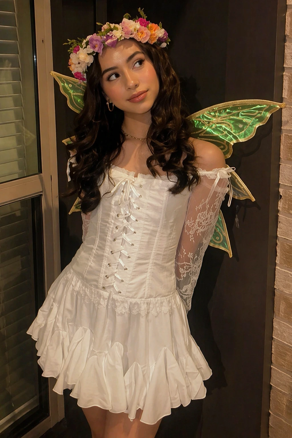 Dark-haired woman in white lace-up fairy dress, flower crown, green wings, posed beside a doorway.