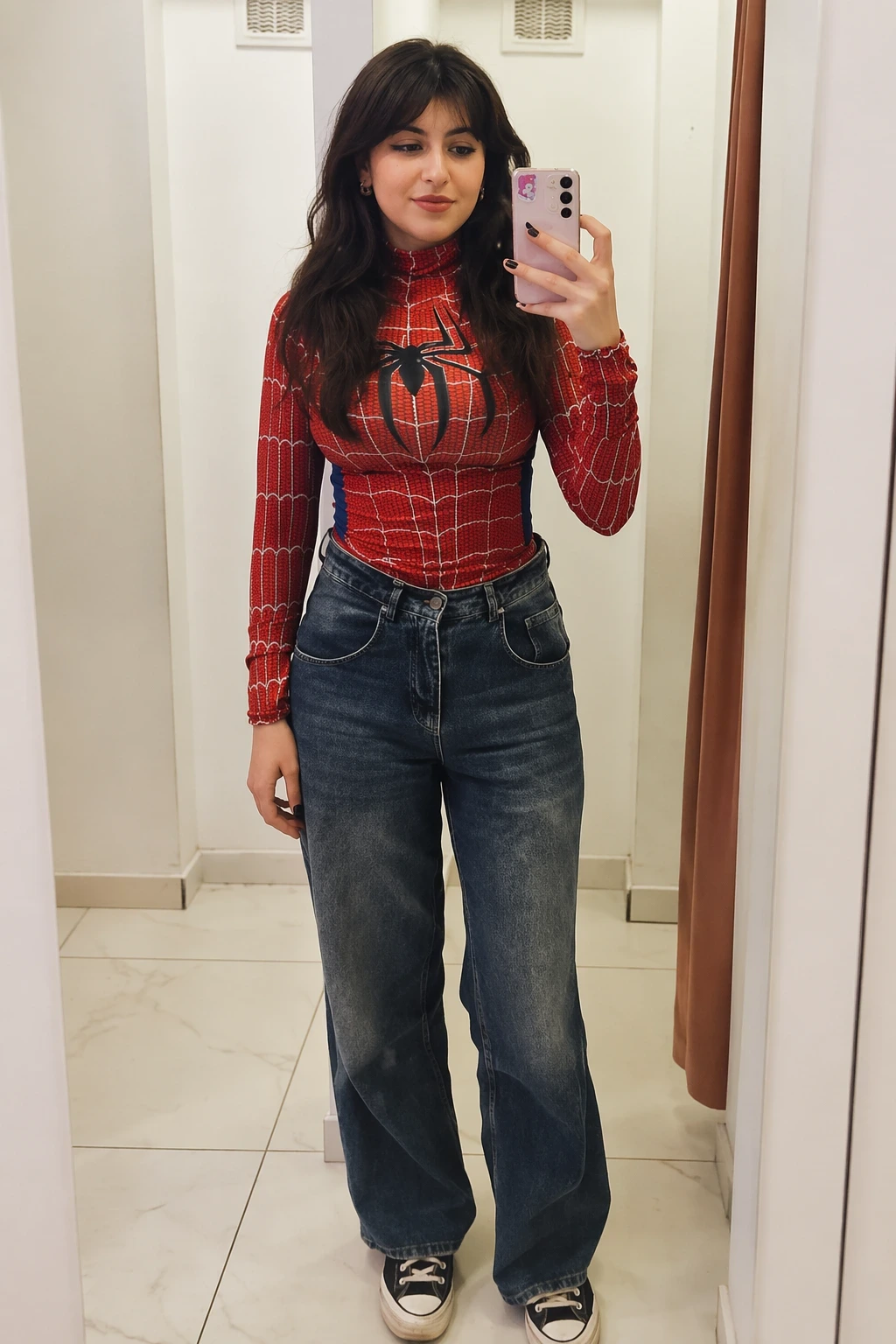 Woman taking a mirror selfie in a red Spider-Man top, loose blue jeans, and black sneakers in a bright fitting room.