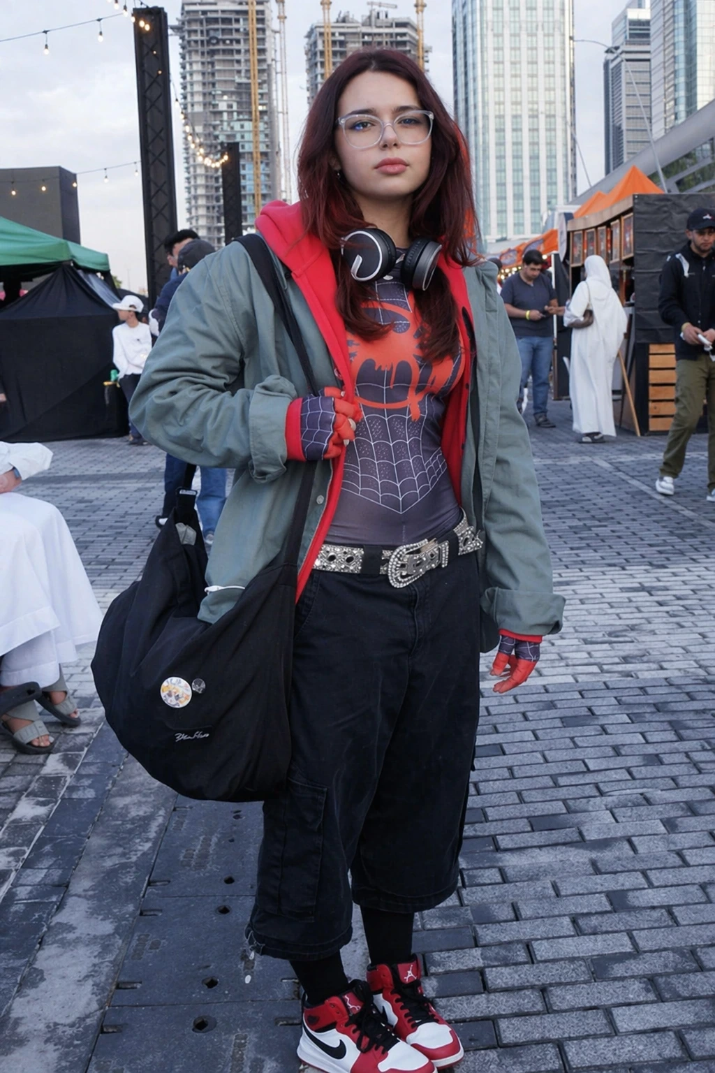 Woman in glasses wearing Spider-Man graphic shirt, green jacket, black shorts, headphones, and red sneakers outdoors.