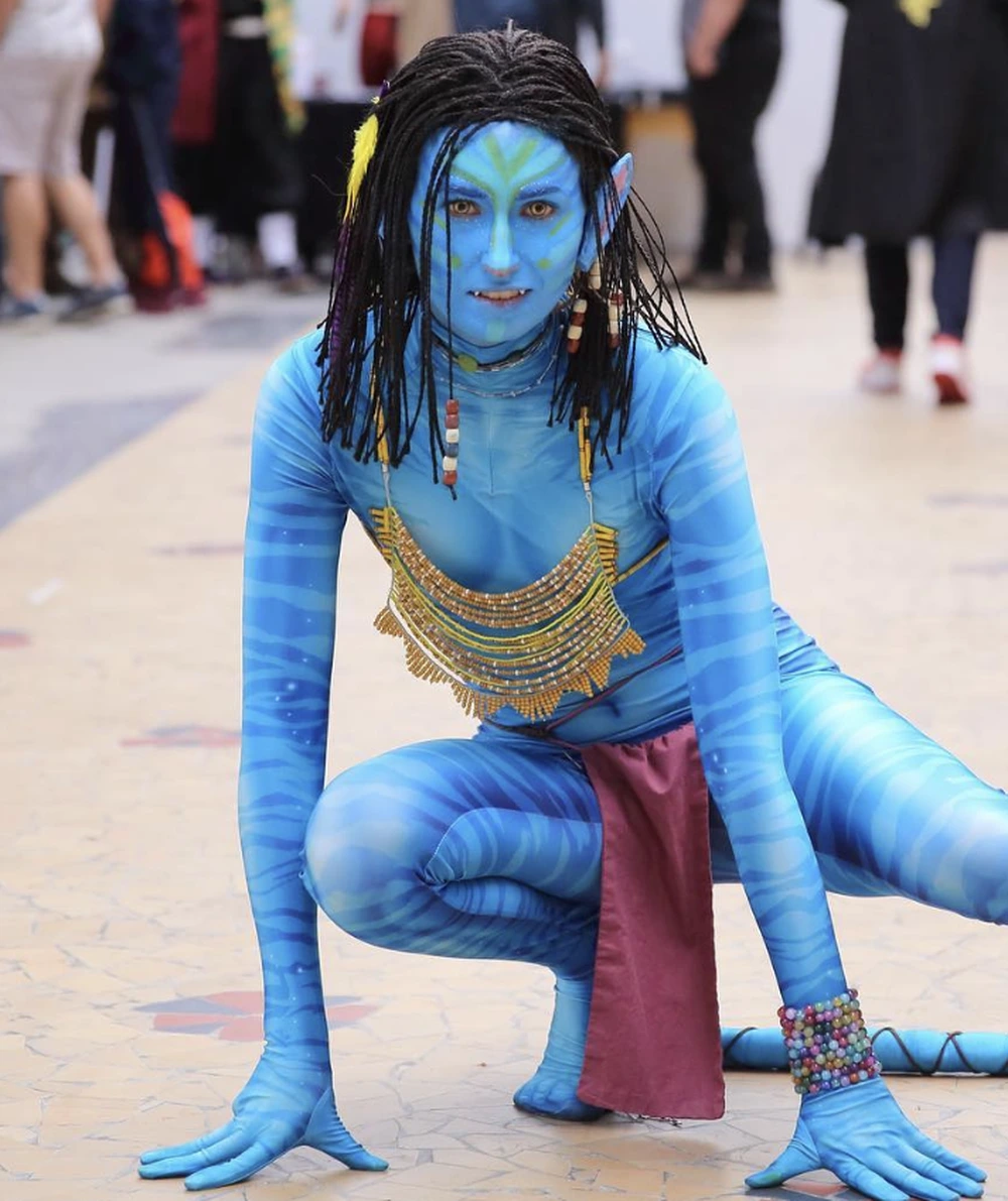 Blue avatar costume in crouched pose, striped body paint, black braids, beaded chest piece, and pink hanging panel.