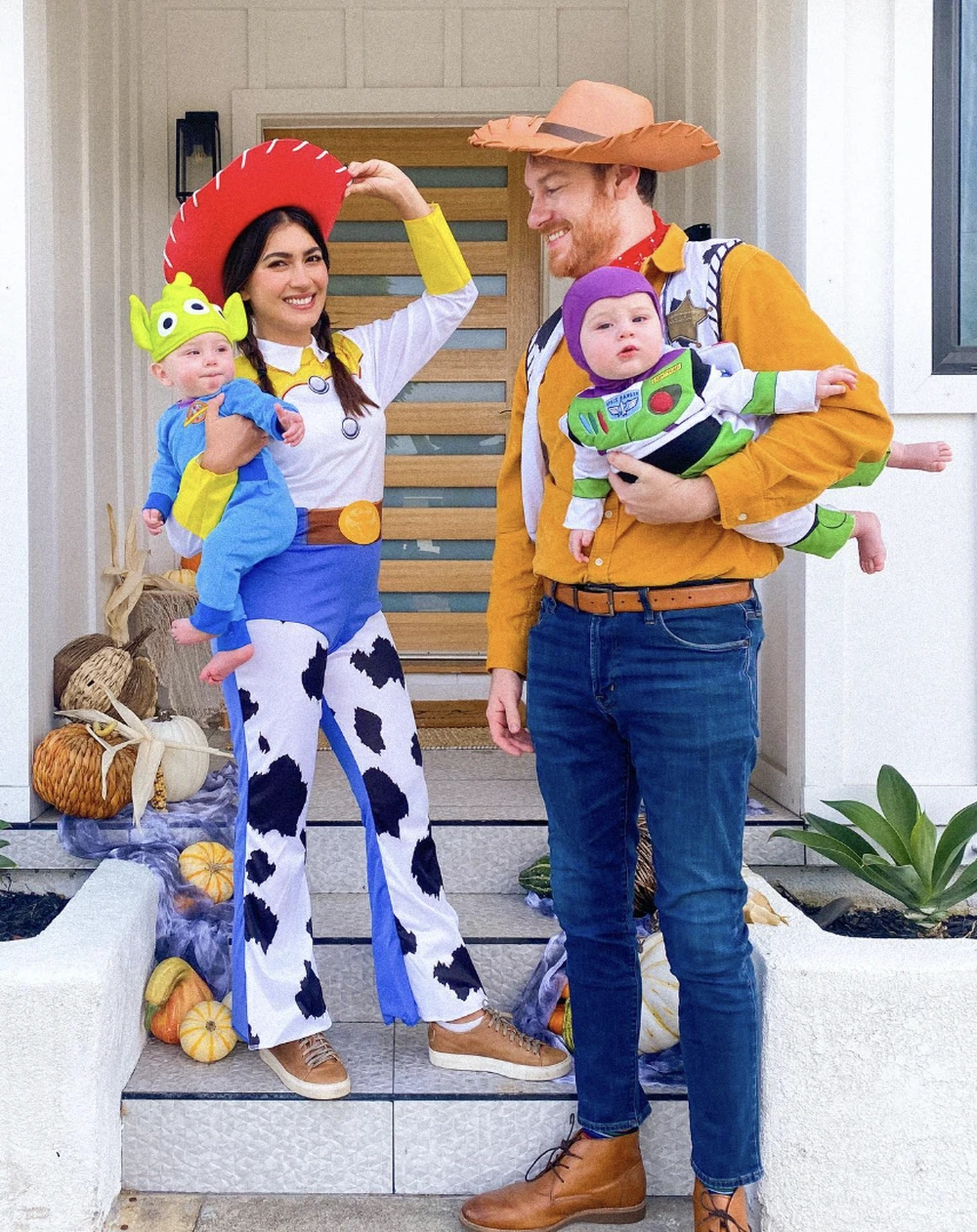 Adults and babies dressed as Woody, Jessie, and Buzz Lightyear on front steps with Halloween pumpkins.