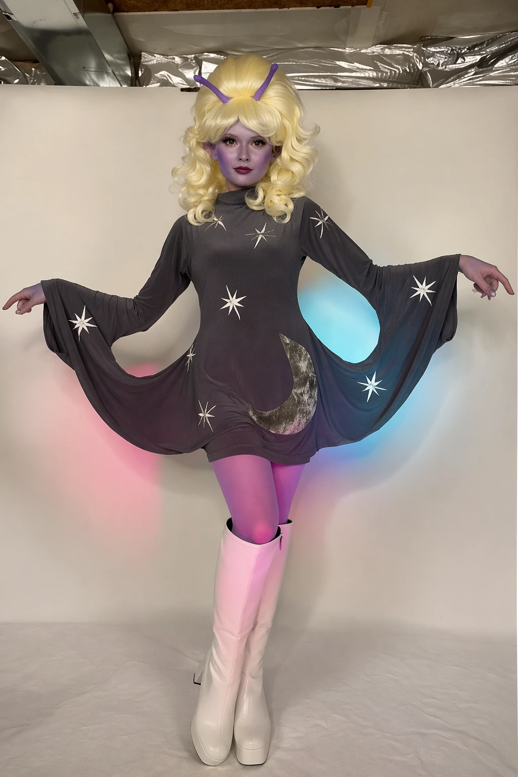 Purple-faced alien costume with blonde curls, antennae, star-patterned dark dress, and white knee-high boots.