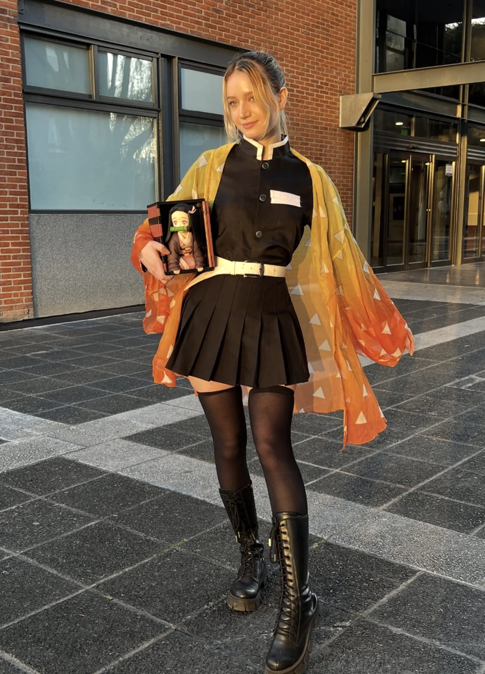 Blonde hair tied back, black uniform, gradient yellow-orange haori, pleated skirt, and tall lace-up boots outdoors.