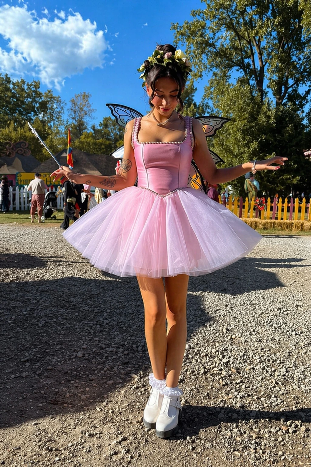 Young woman in pink tutu dress, floral crown, butterfly wings, and white platform shoes at outdoor festival.