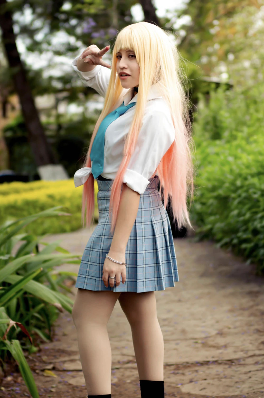 Blonde wig with pink ends, white shirt, blue tie, plaid skirt, and black socks on a garden path.