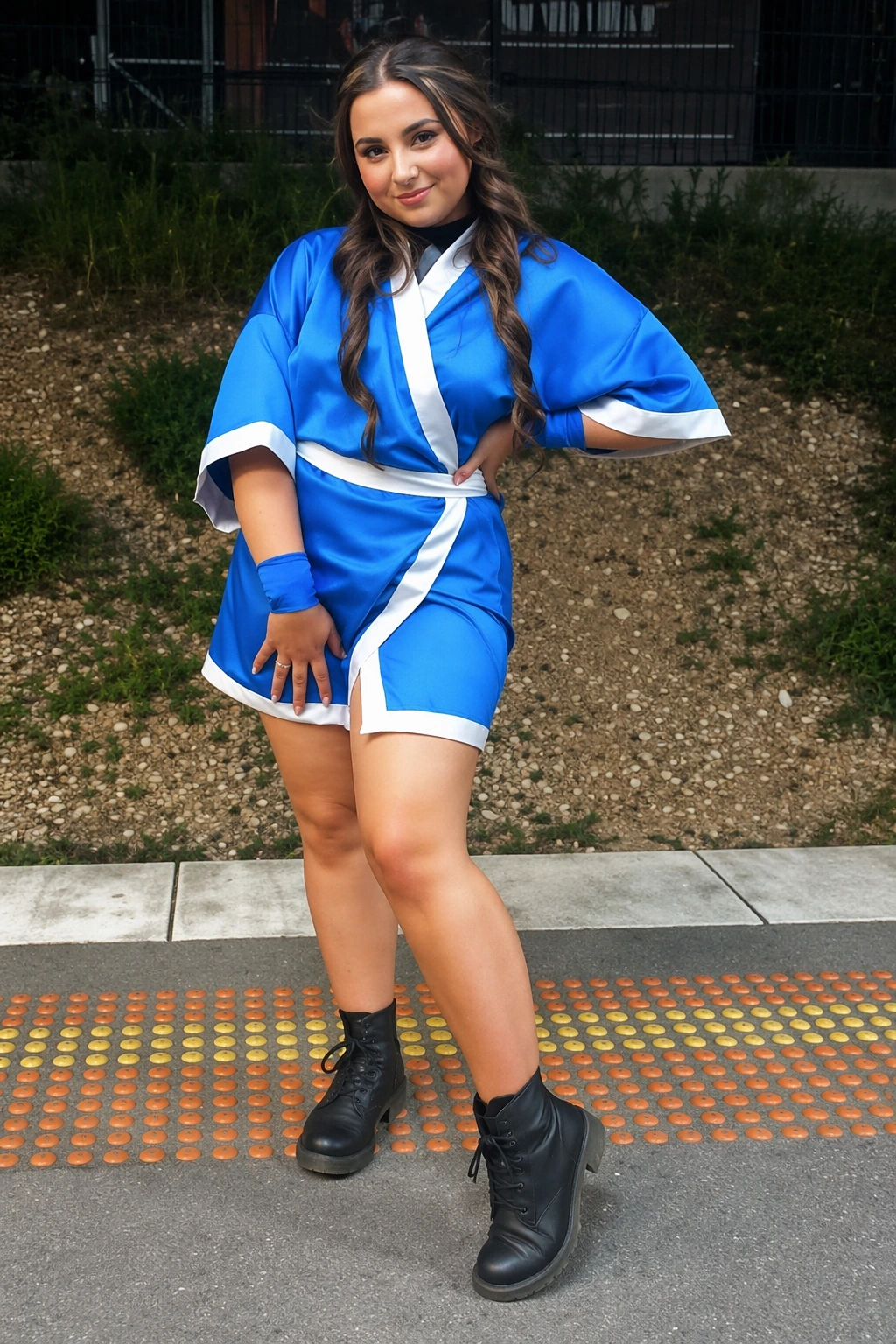 Smiling woman stands outdoors in bright blue robe-style costume, black ankle boots, and loose wavy hair.