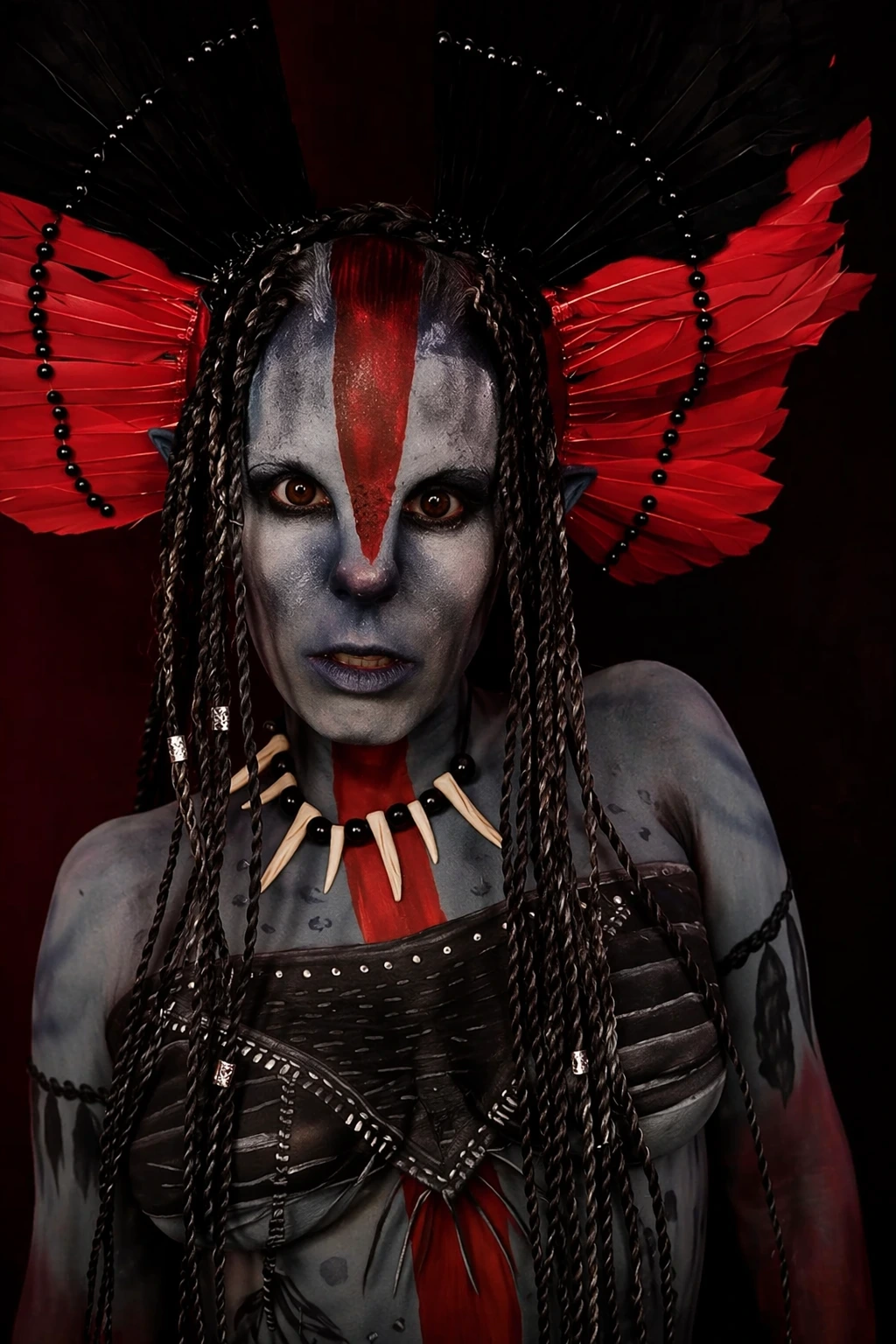 Tight portrait of blue-gray avatar makeup, braided hair, red-and-black headdress, bone necklace, and intense eye makeup.