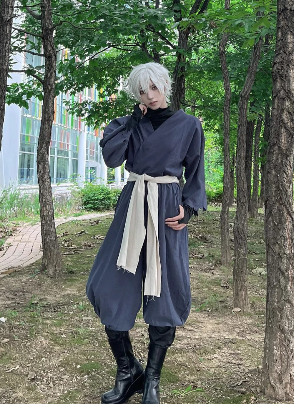 White-haired cosplayer in loose charcoal robe, cream sash, and black boots standing among trees.