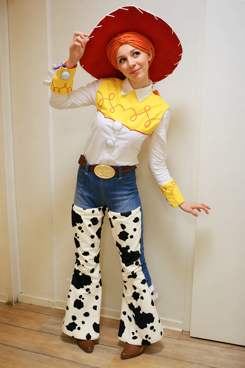 Woman dressed as Jessie in a white shirt, yellow yoke, cow-print chaps, blue jeans, and oversized red hat.