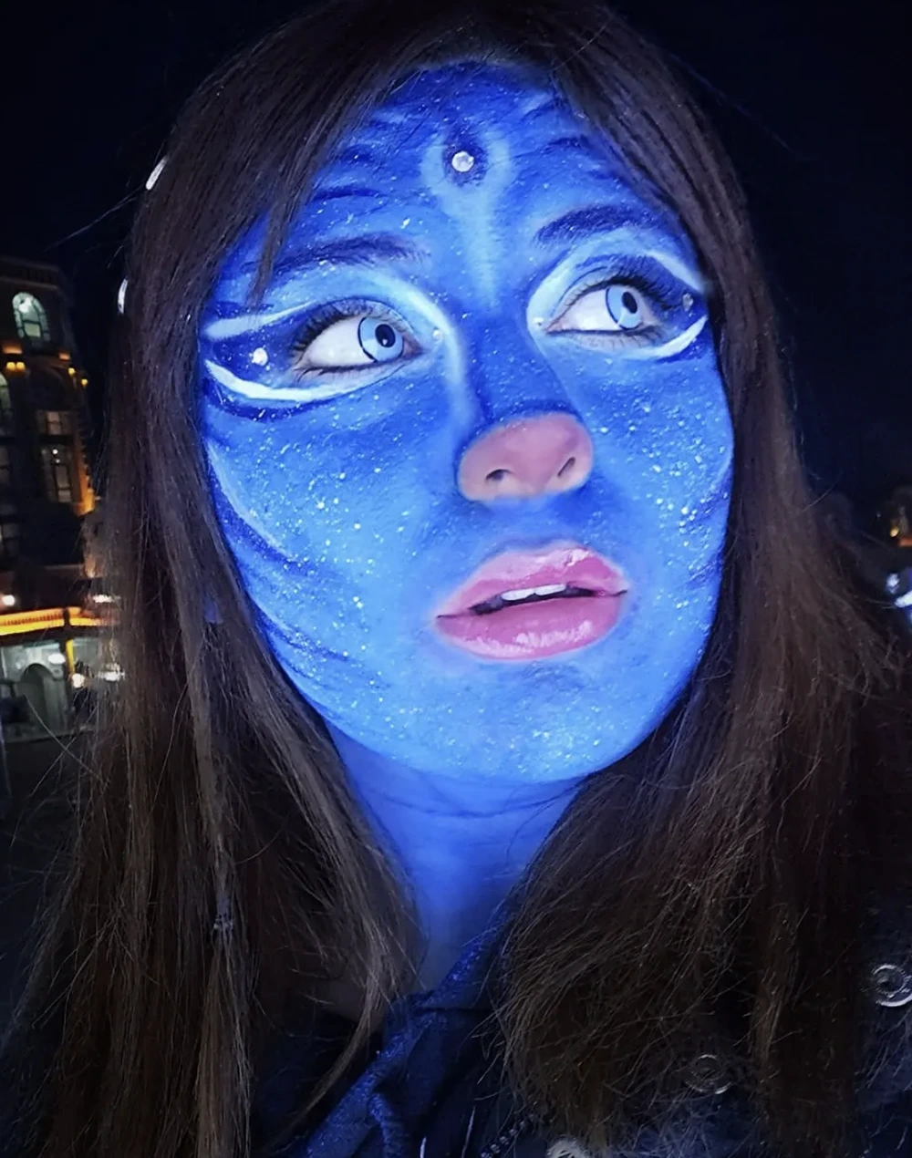 Nighttime close-up of blue-painted avatar face with white eye accents, sparkly finish, and long dark hair.