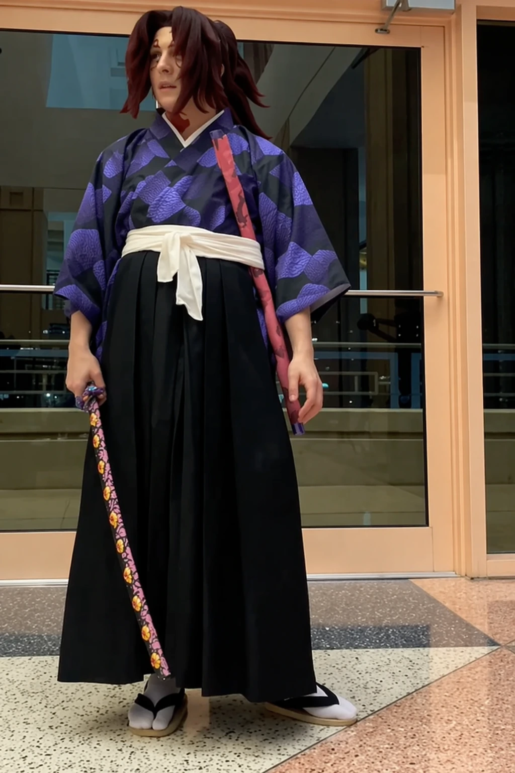 Cosplayer in dark patterned kimono, white sash, wide black hakama pants, sandals, and floral-handled sword prop.