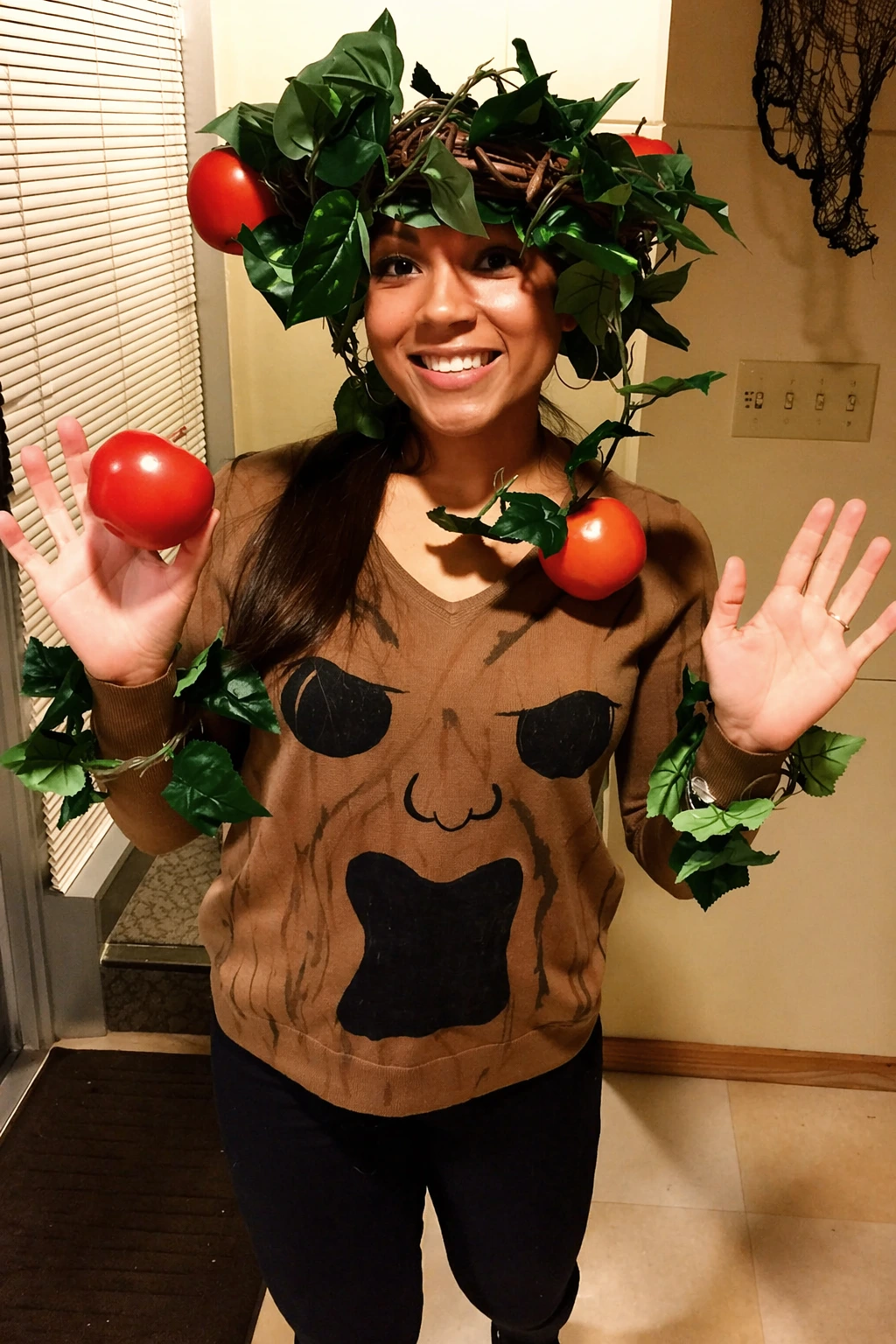Woman wears a brown tree-themed outfit with green leaves and red tomato-like decorations indoors.