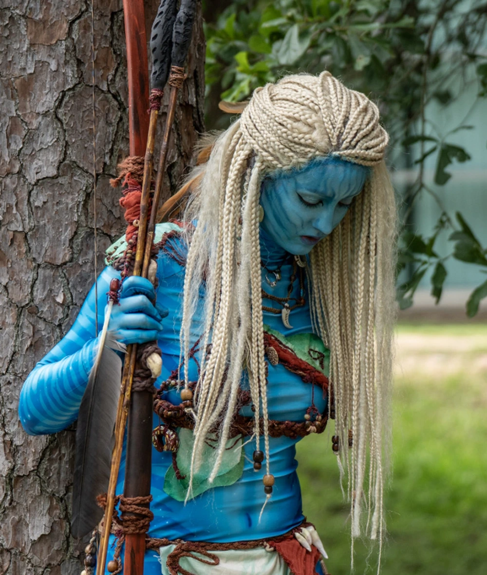 Blue-skinned avatar costume with long platinum braids, holding a spear beside a tree in outdoor daylight.