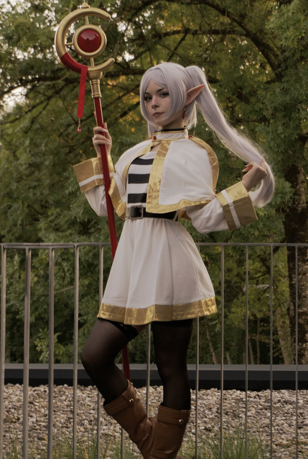 Silver-haired elf cosplayer wearing white robe with gold trim, black tights, and holding a red-gold staff outdoors.