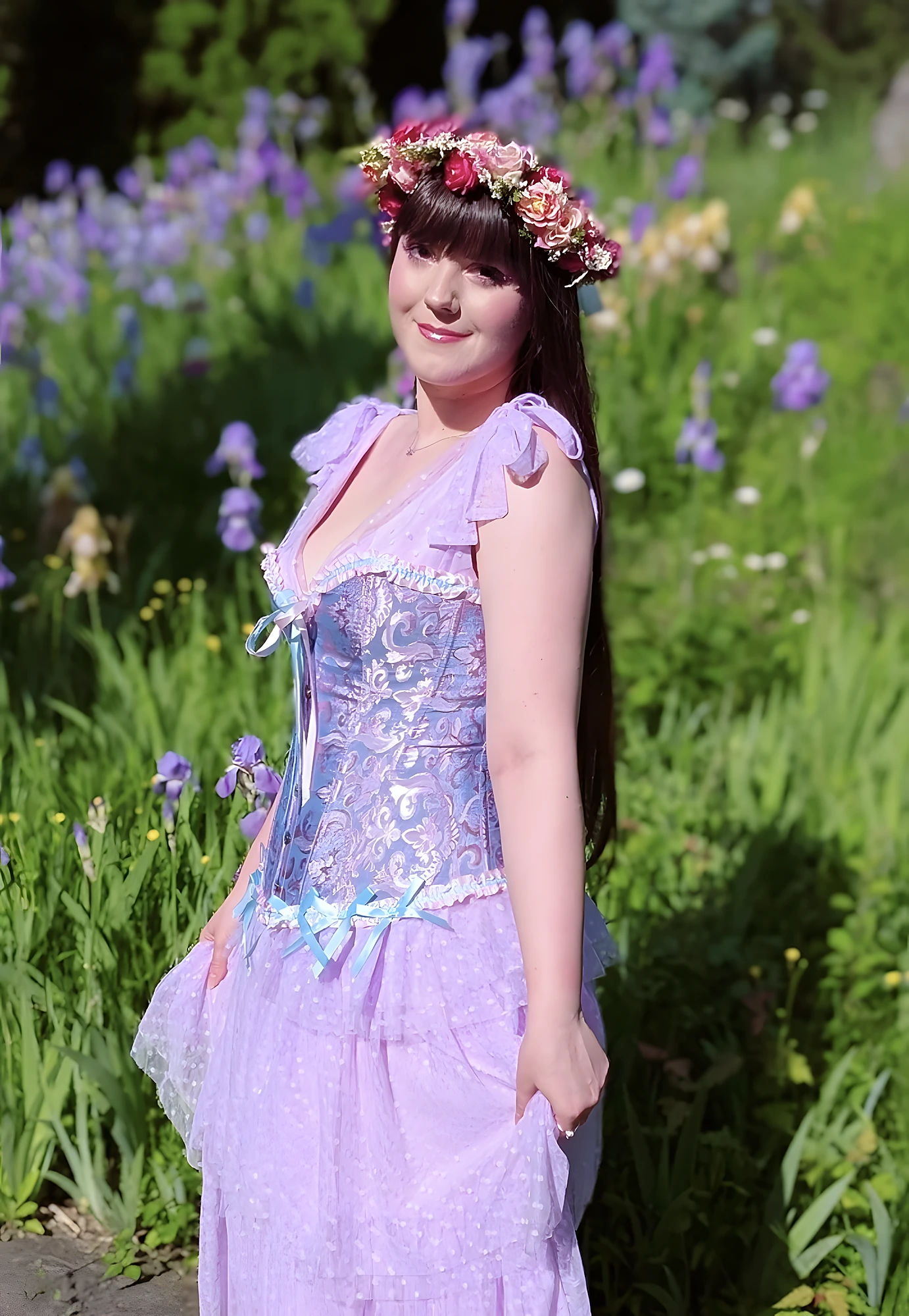 Woman wearing lilac floral corset dress and flower crown, posed in sunlit garden with purple blossoms.