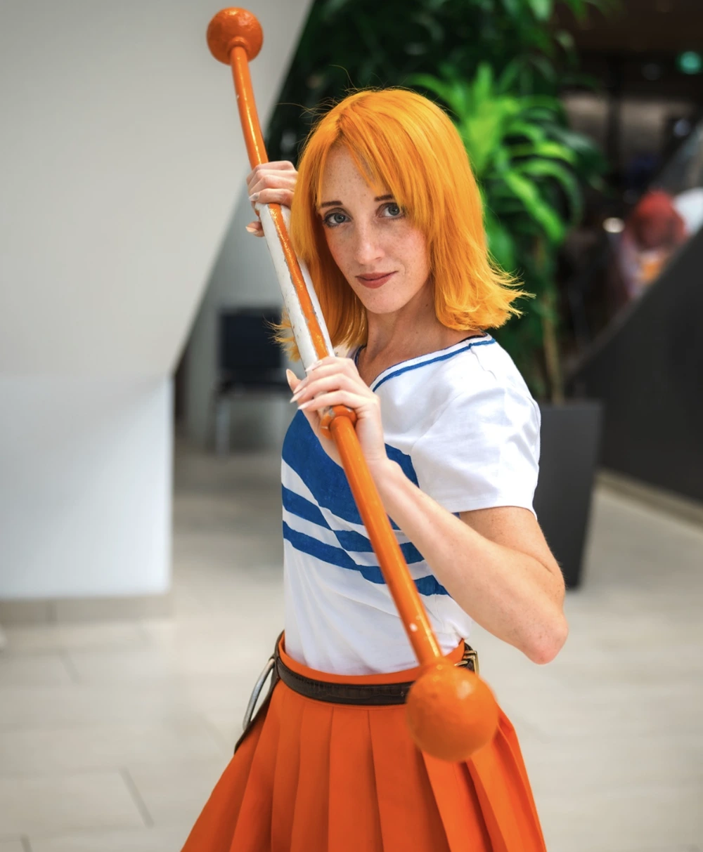 Orange-haired cosplayer in white-and-blue top, orange pleated skirt, holding a long orange staff indoors.