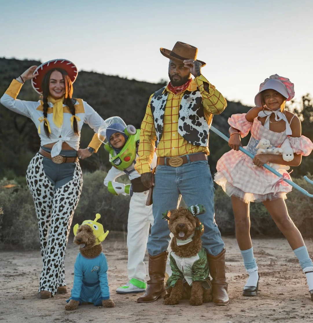 Four people and two dogs wear coordinated Toy Story outfits, including Woody, Buzz, Jessie, and alien-inspired costumes outdoors.