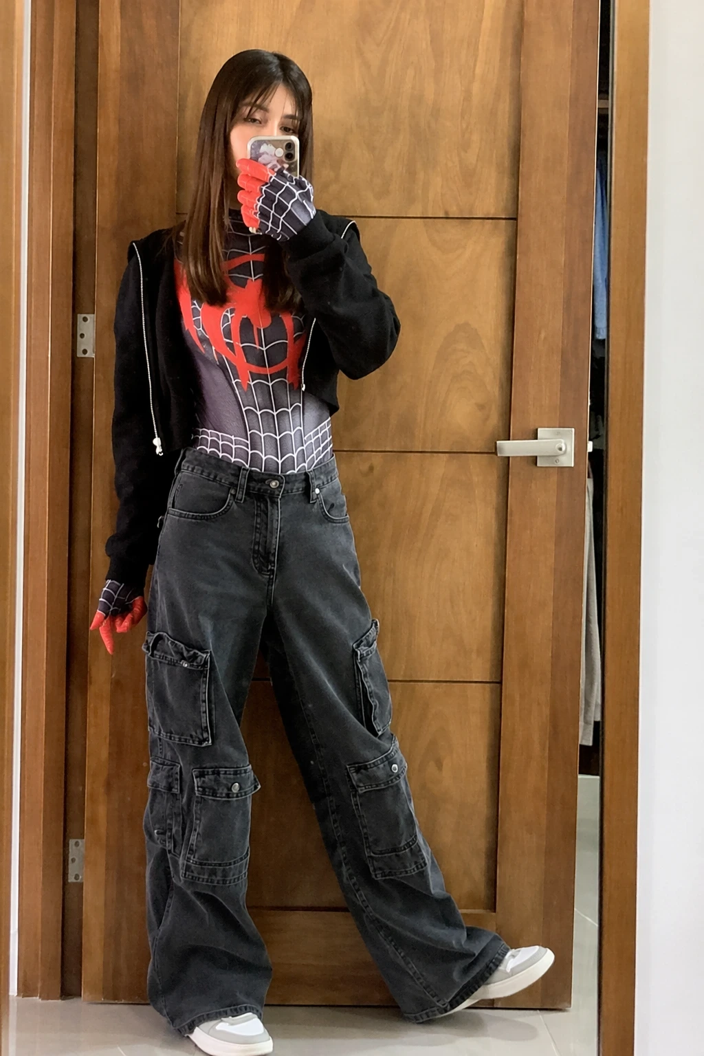 Person posing in a black hoodie, red-and-gray Spider-Man bodysuit, cargo jeans, gloves, and white sneakers indoors.